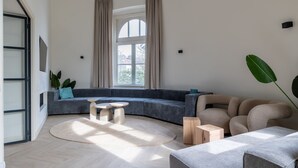 Un salon moderne avec un canapé d’angle aux lignes courbes, une table basse ronde et une grande fenêtre agrémentée de rideaux.