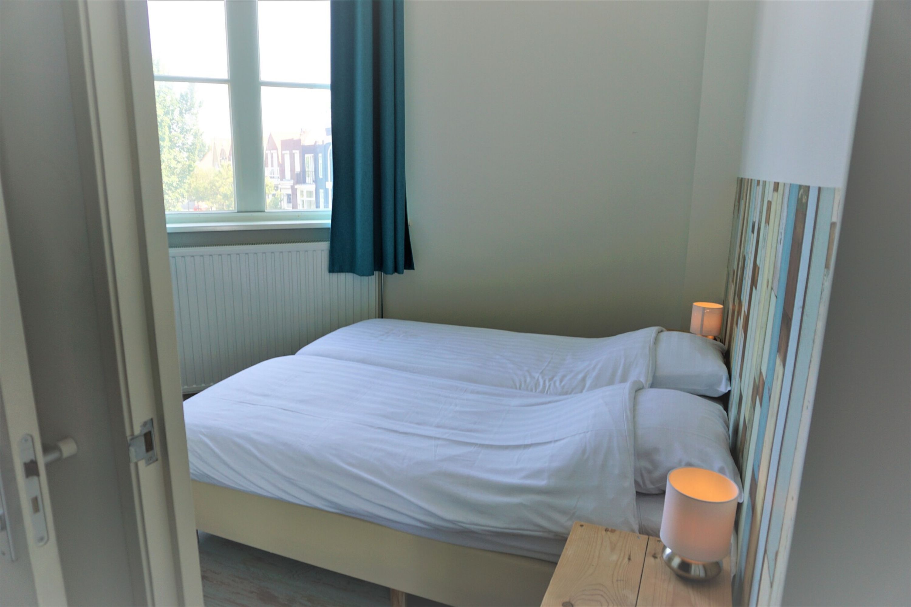 A bedroom with a bed, a bedside table with a lamp, a window with curtains, and a radiator.