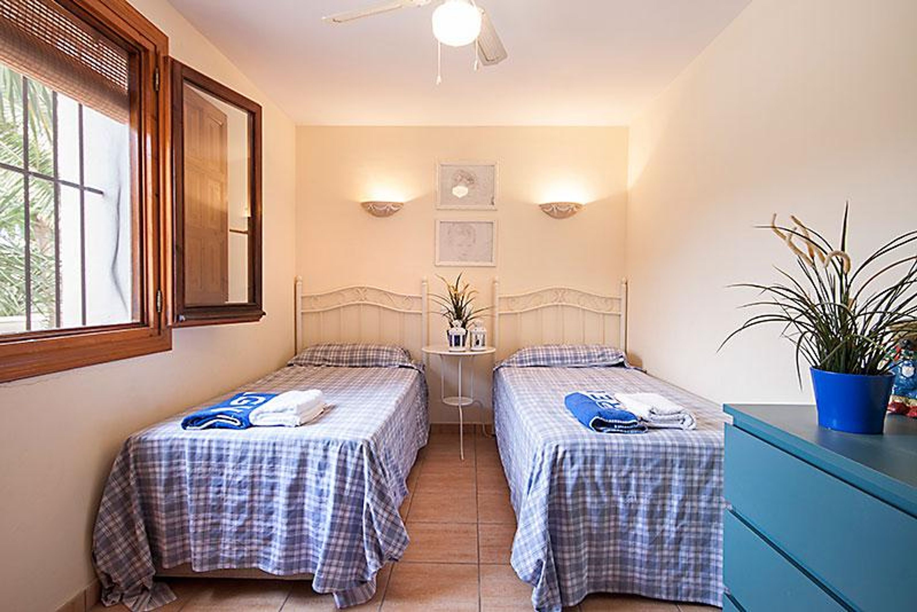 A room with two single beds, a small table with a plant, and a ceiling fan.