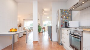 Une cuisine moderne avec des armoires blanches, un réfrigérateur en acier inoxydable et un four encastré. On aperçoit, au fond, un coin repas avec une table et des chaises.