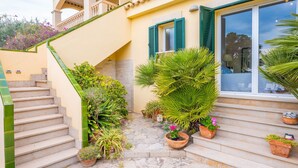 Une maison avec un escalier menant à un balcon, entourée de plantes en pot et d’un chemin de pierre.