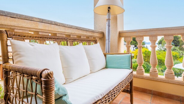 Un canapé en osier agrémenté de coussins blancs et turquoise, placé sur un balcon extérieur doté d’une balustrade en pierre et d’un lampadaire.