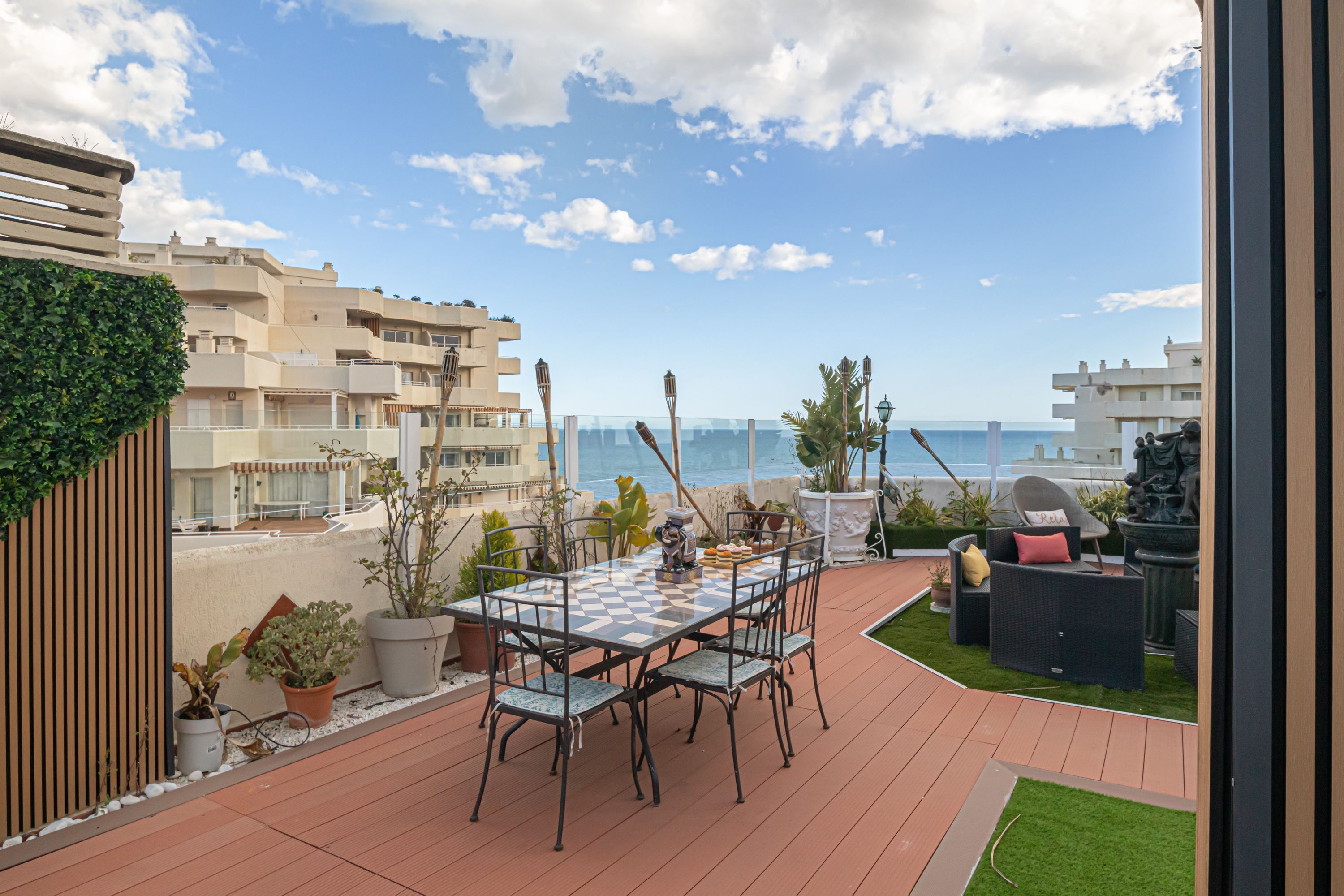 A terrace with a checkered table and chairs, potted plants, and a view of the sea.