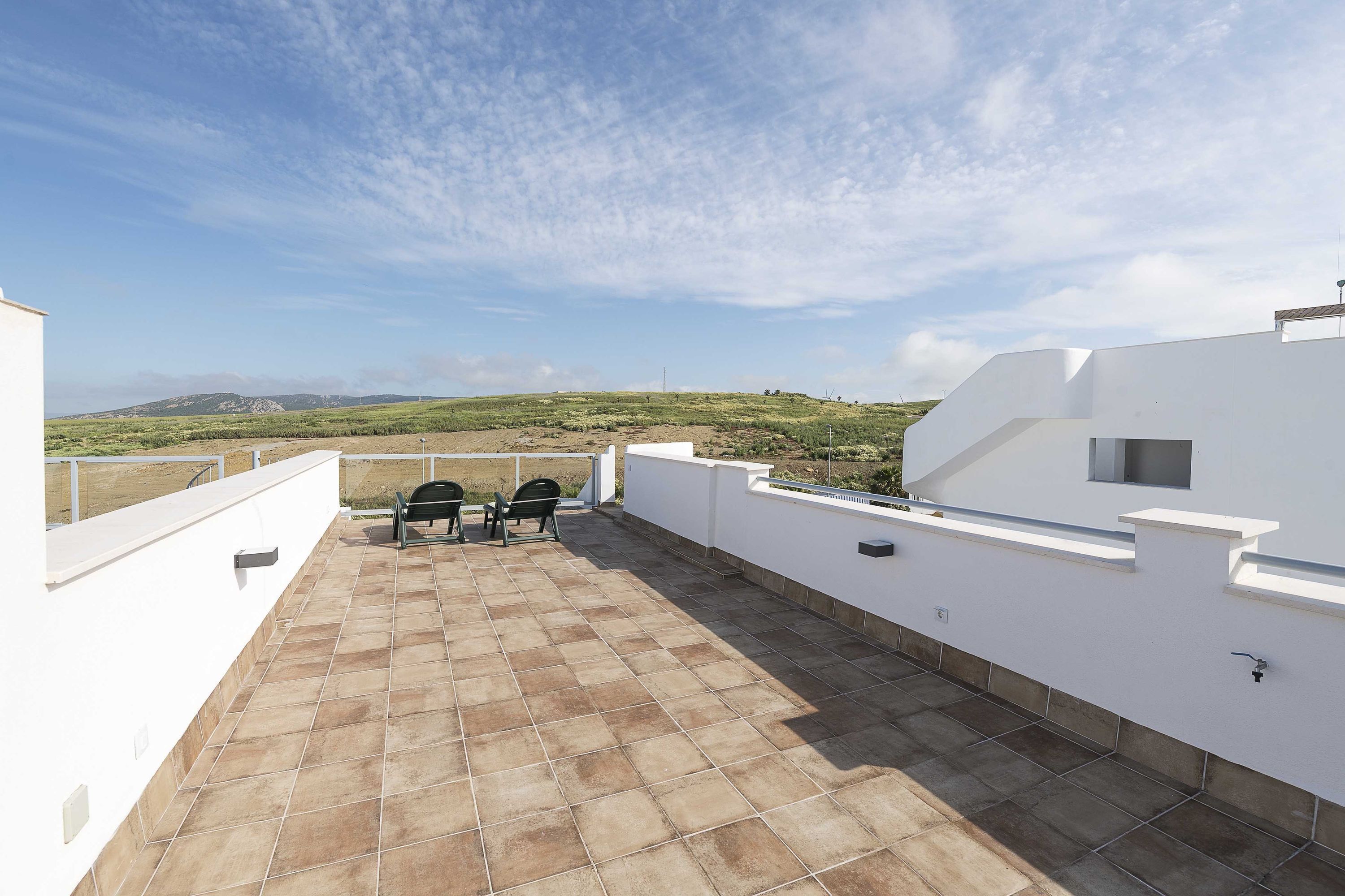 Terraza en la azotea con vistas al campo, que incluye un edificio blanco, suelo de baldosas y dos sillas.