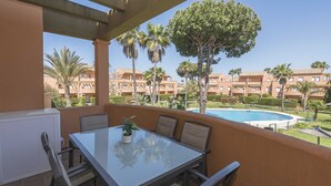 A balcony with a glass table and chairs, overlooking a swimming pool and a landscaped garden with palm trees. - Novo Golf Chiclana in Chiclana de la Frontera (Chiclana de la Frontera)