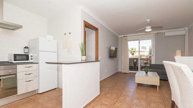 A modern kitchen with white cabinetry, a microwave, and a refrigerator. There is a small island with a plant, and a dining area with a sofa and a coffee table. - Novo Golf Chiclana in Chiclana de la Frontera (Chiclana de la Frontera)