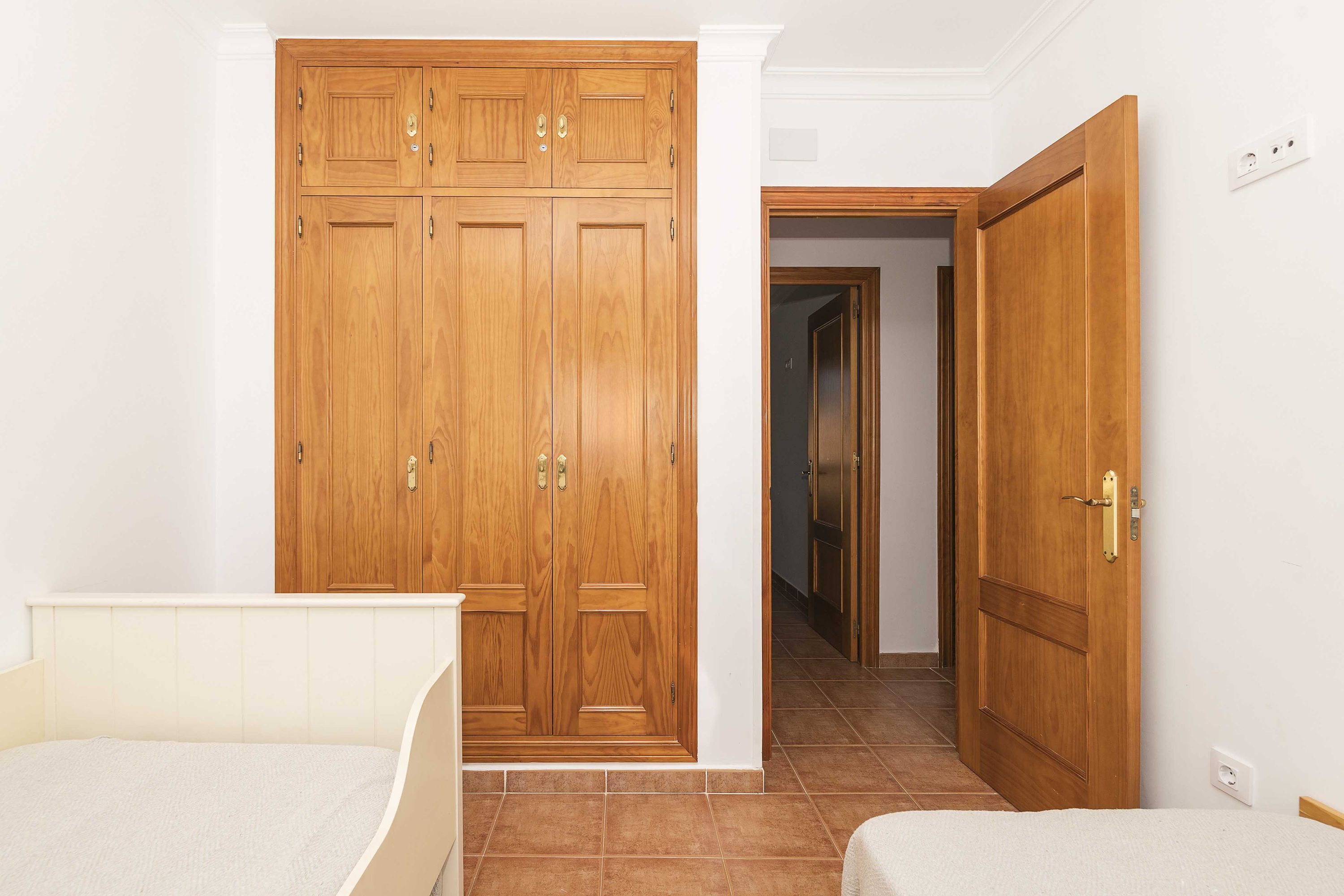 A bedroom with a wooden wardrobe, a bed, and a door leading to another room.