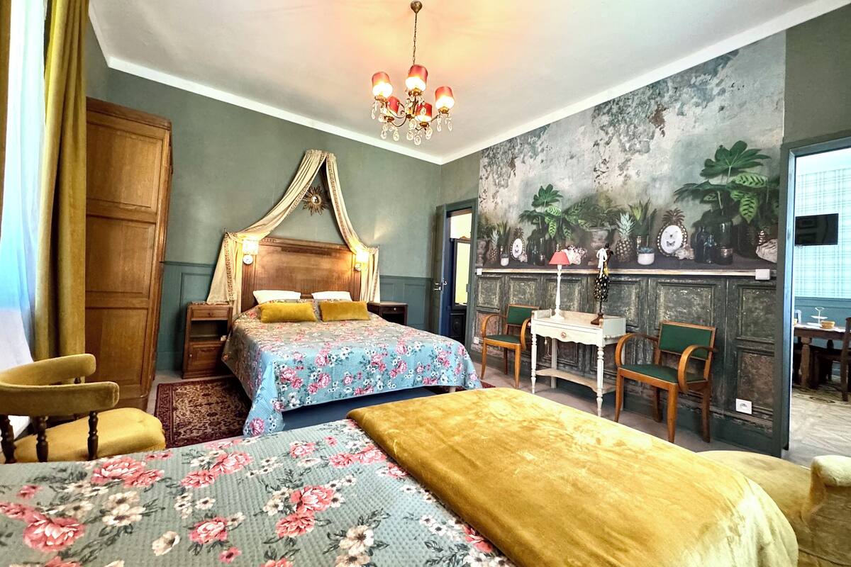 A bedroom with a canopy bed, a floral bedspread, a green chair, and a mural of tropical plants.