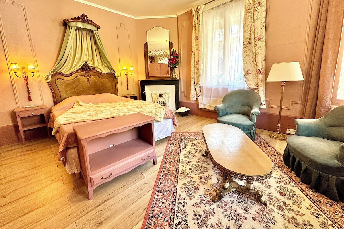 A bedroom with a bed, a chair, a nightstand, a lamp, a mirror, and a carpet.