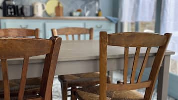 A dining area with wooden chairs and a table, a kitchen in the background with blue cabinets and shelves, and a patterned floor.