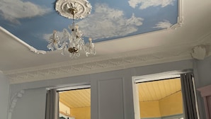Ein Zimmer mit einer Decke, die mit Wolken bemalt ist, einer Chandelüster und einem Blick durch eine offene Tür in ein weiteres Zimmer mit einem Fenster.
