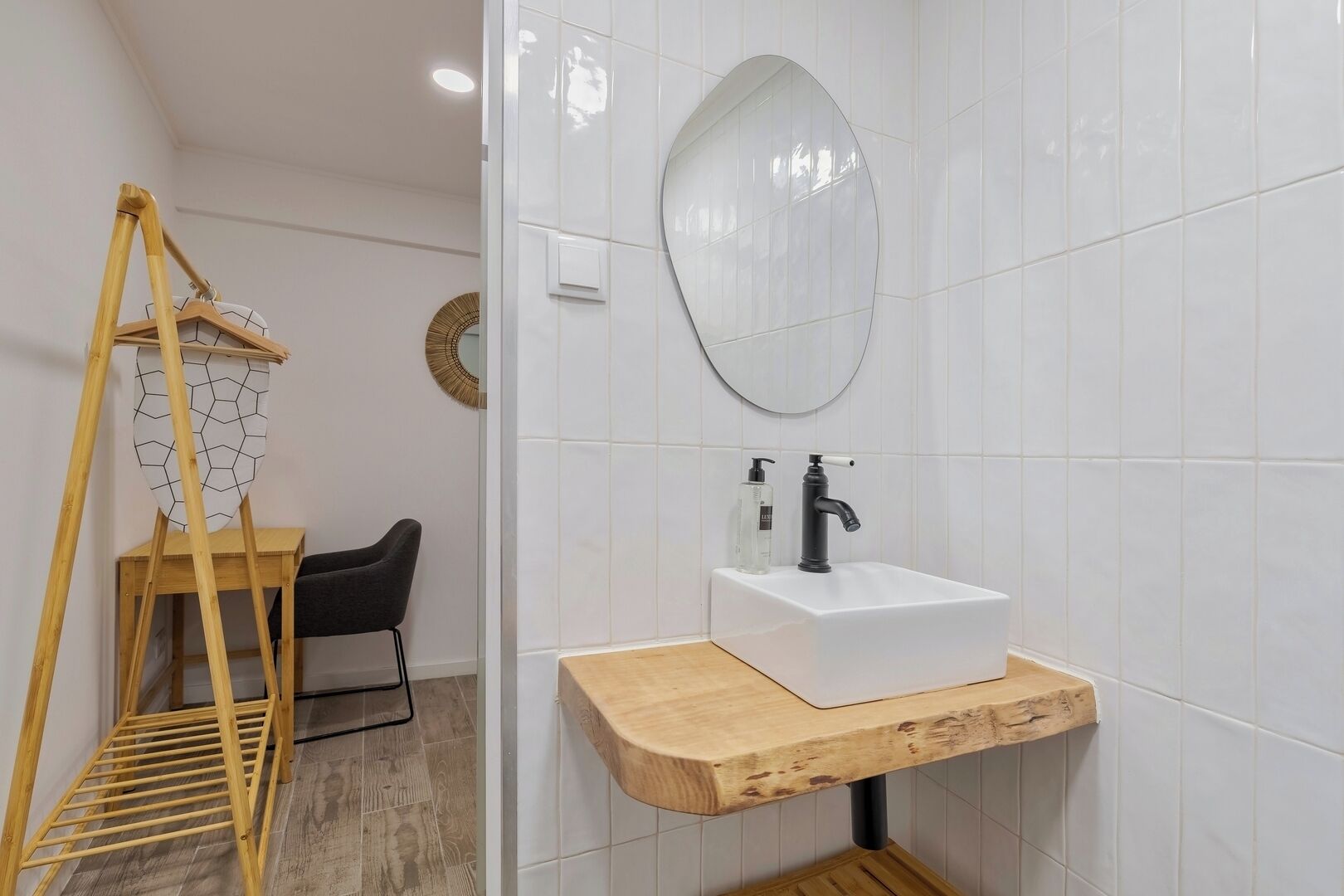 Een badkamer met een houten droogrek, een zwarte stoel, een witte wastafel met houten blad en een ronde spiegel aan de muur.