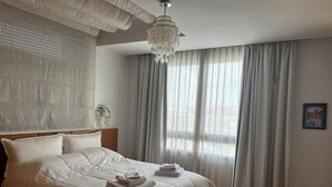 Une chambre avec un grand lit, un lustre, une vue sur la ville à travers de fines rideaux, et un tableau accroché au mur.