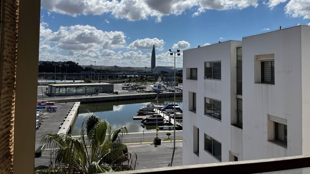 Une vue d’une marina avec des bateaux amarrés, un bâtiment moderne et un ciel dégagé avec quelques nuages.