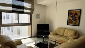 Un salon moderne doté d’une grande fenêtre, d’un téléviseur à écran plat, d’un canapé beige et d’une table basse en verre.
