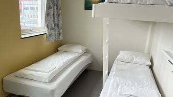 A dormitory room with two single beds, a window with curtains, a painting on the wall, and a view of buildings outside.