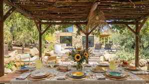 An outdoor dining area with a pergola, stone seating, and a view of a modern house.
