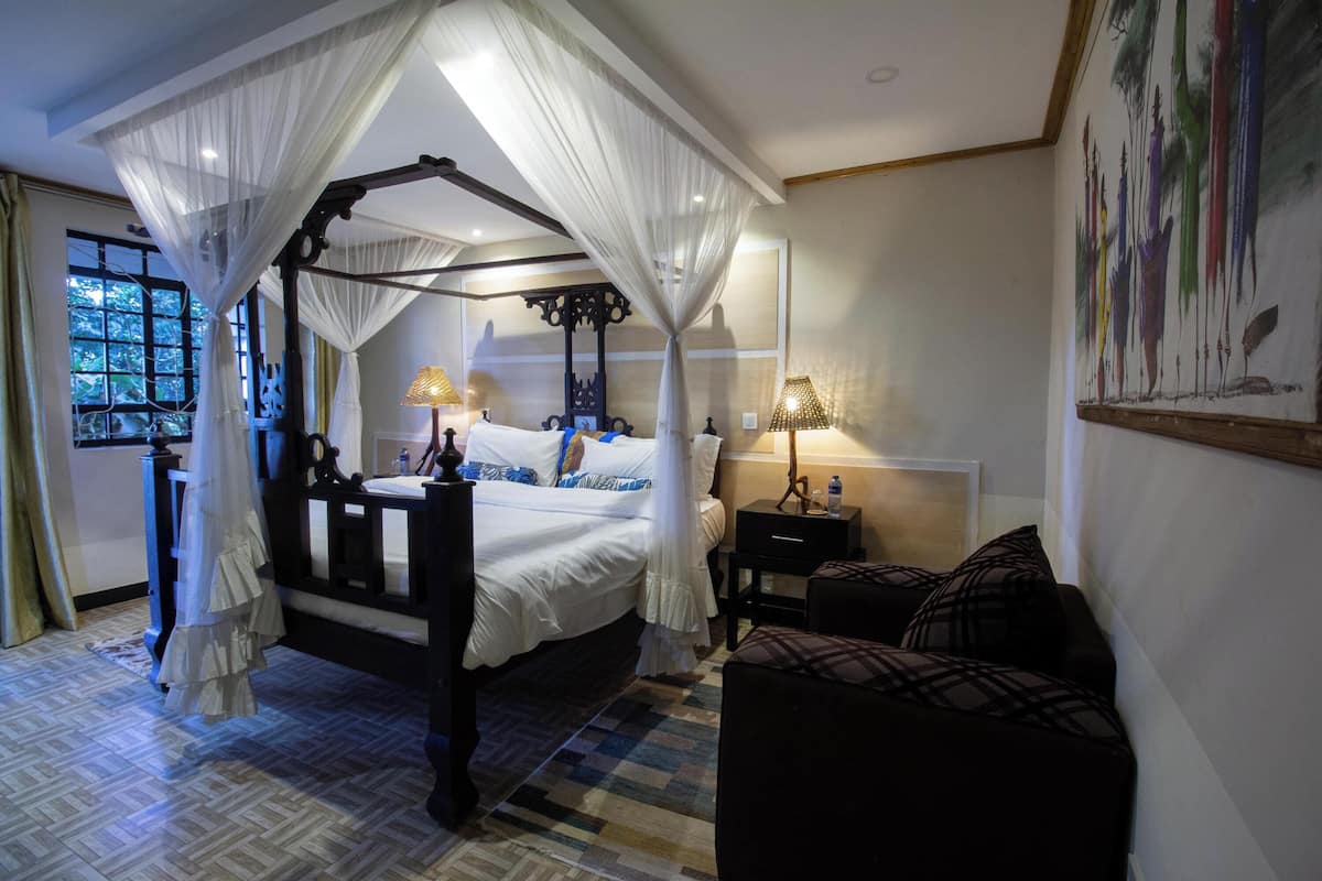 A four-poster canopy bed with white bedding, a bedside table with a lamp, and a view of the outdoors through a window.