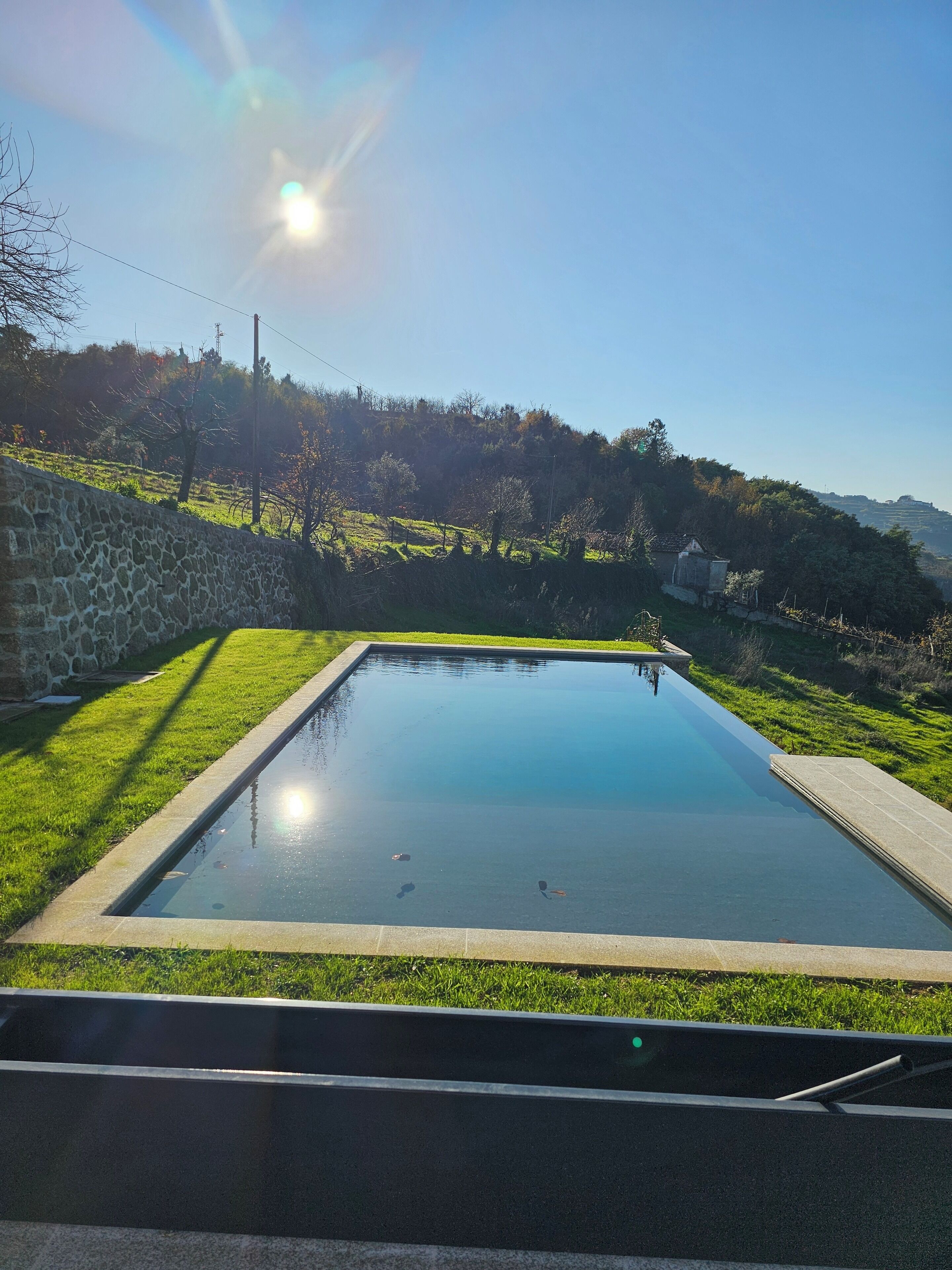 A rectangular swimming pool with a stone wall on one side and a grassy area on the other.
