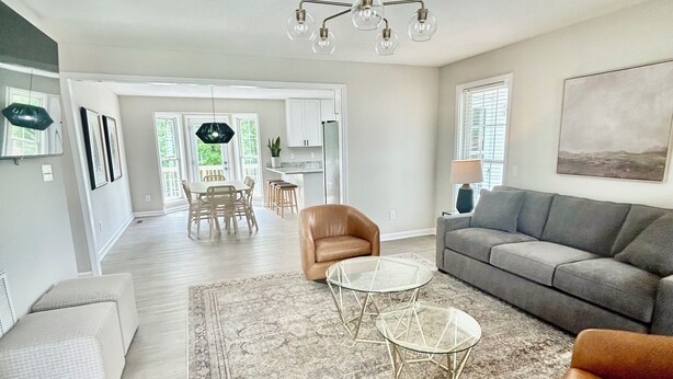 A spacious living room with a grey sofa, a brown leather chair, and two glass coffee tables. There is a large rug on the floor, and the room has a modern chandelier.