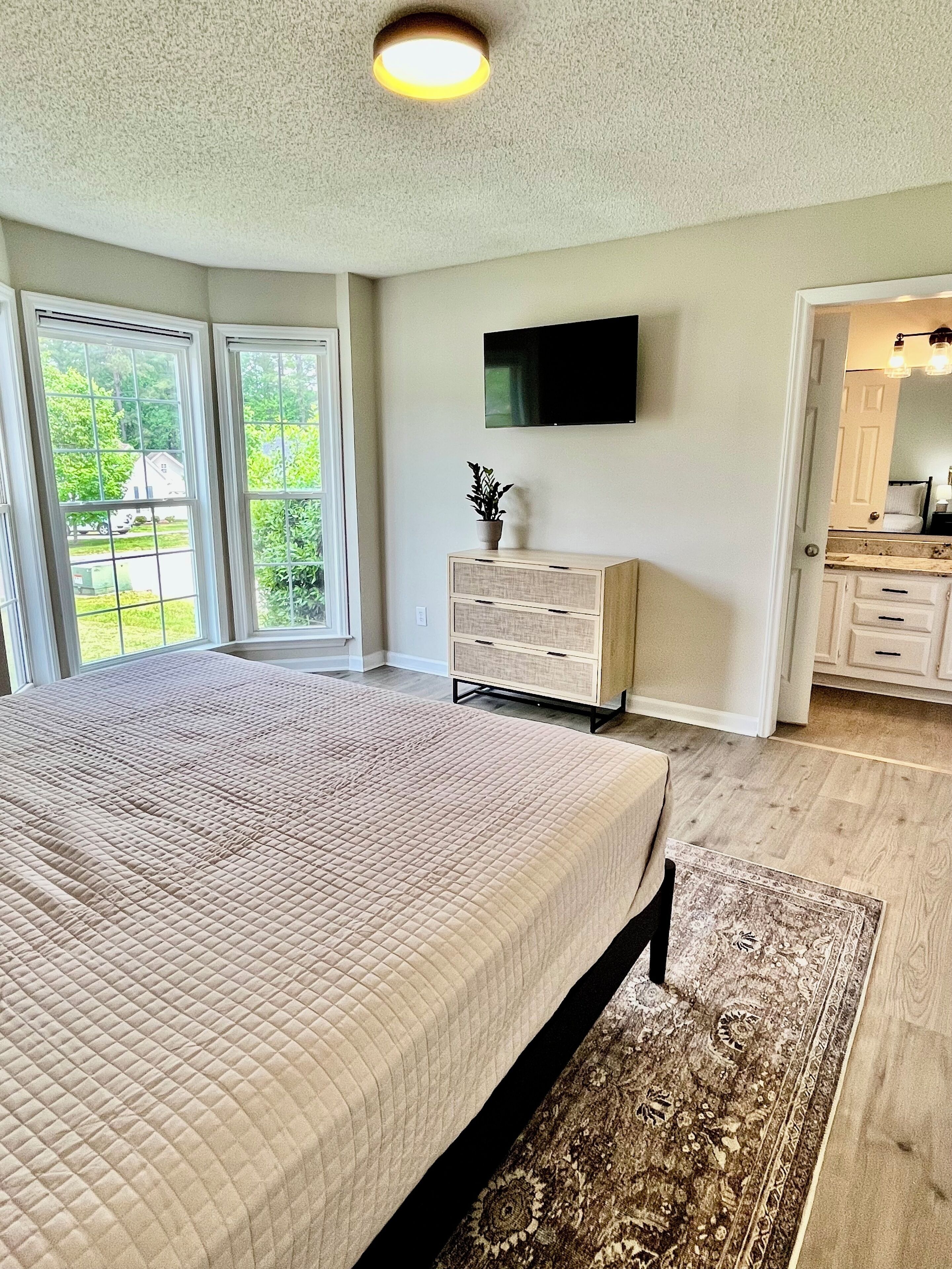 A bedroom with a large bed, a television on the wall, and a view of the outdoors through the windows.