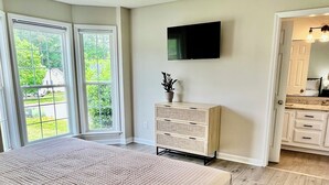 A bedroom with a large bed, a television on the wall, and a view of the outdoors through the windows.