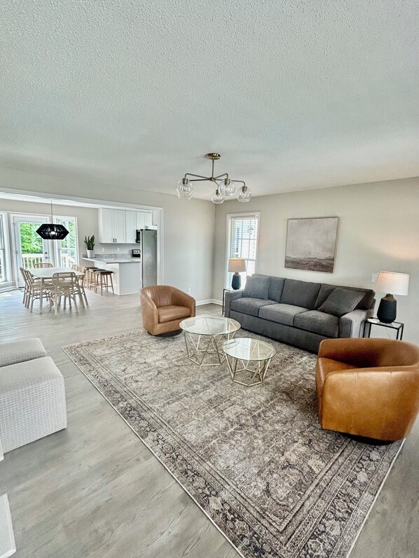 A spacious living room with a grey sofa, two round coffee tables, a brown armchair, and a large rug. - 5BR/3BA Sleeps 12 NEW kitchen, bathrooms, mattresses, linens, furniture, firepit (Raleigh)