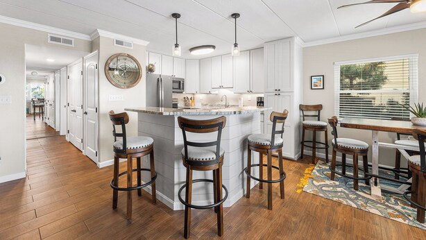 A modern kitchen with white cabinetry, a central island with bar stools, a large clock, and a ceiling fan. - Sea Winds North 112, North OC, Close to the Beach (Ocean City)