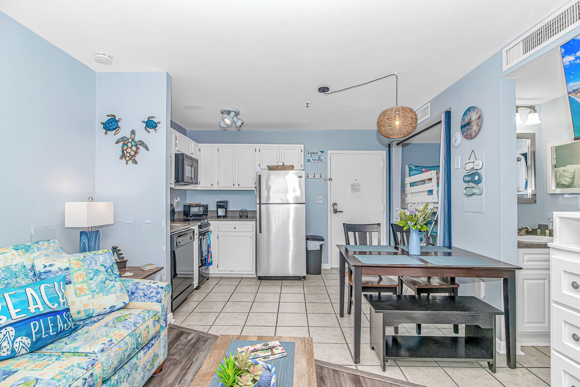 A compact kitchen with white cabinets, a refrigerator, a microwave, and a stove. There's a dining area with a table and chairs, and a sofa with a beach-themed pillow.