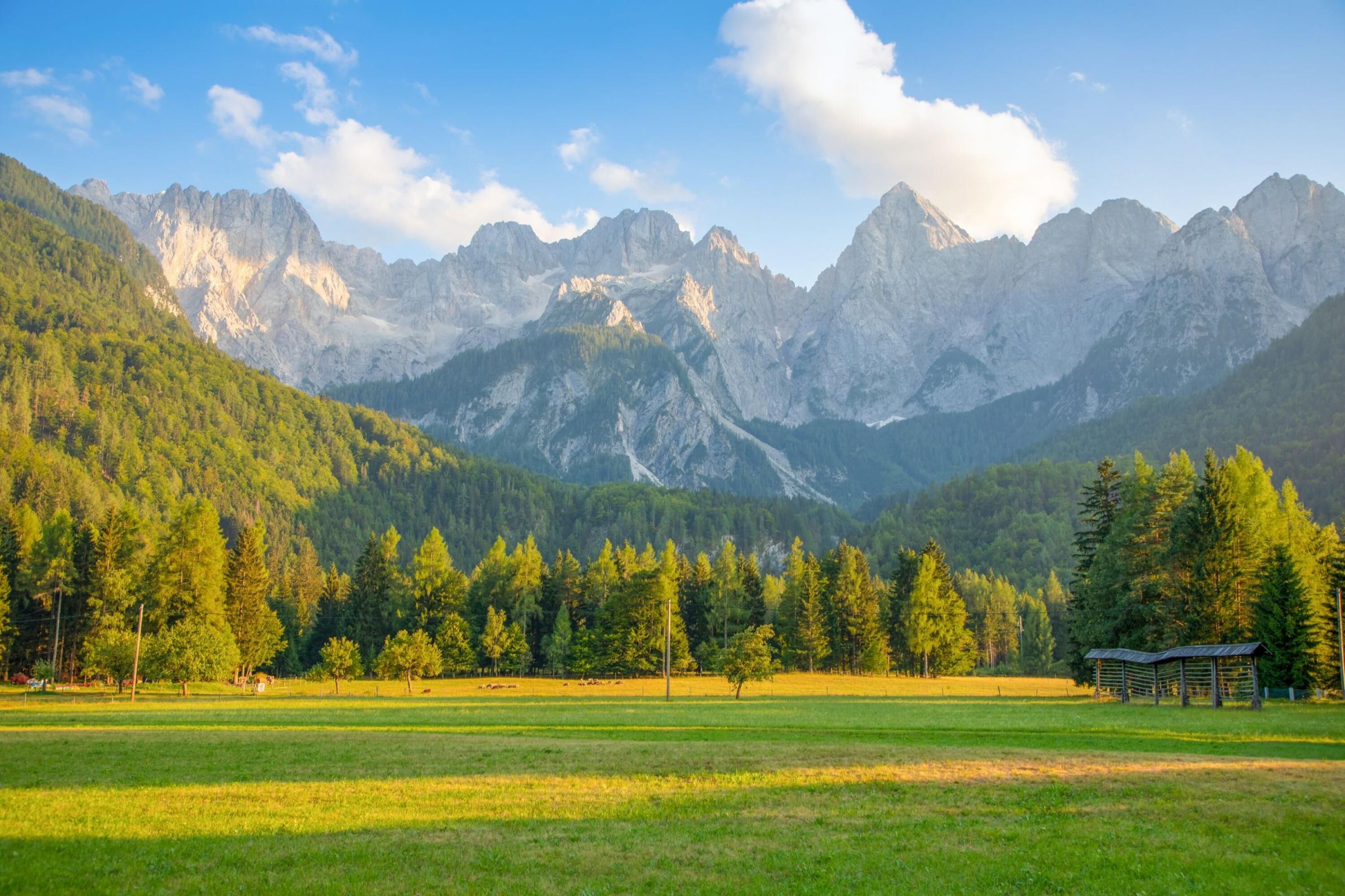 A mountainous landscape with a clear sky, a grassy field, and a small structure in the foreground.