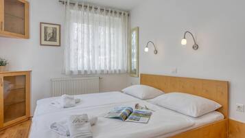 A neatly made bed with white linens, a bedside table with a lamp, and a window with sheer curtains.
