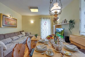 Un salón luminoso con un sofá, una mesa de comedor preparada para dos personas y un televisor que muestra un paisaje panorámico.