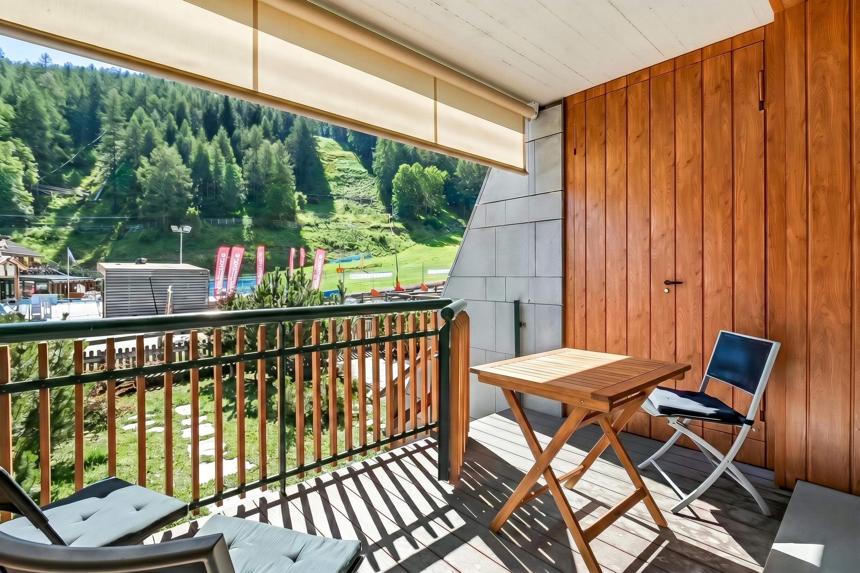 A balcony with a view of a forested hillside, a wooden table, and a chair.