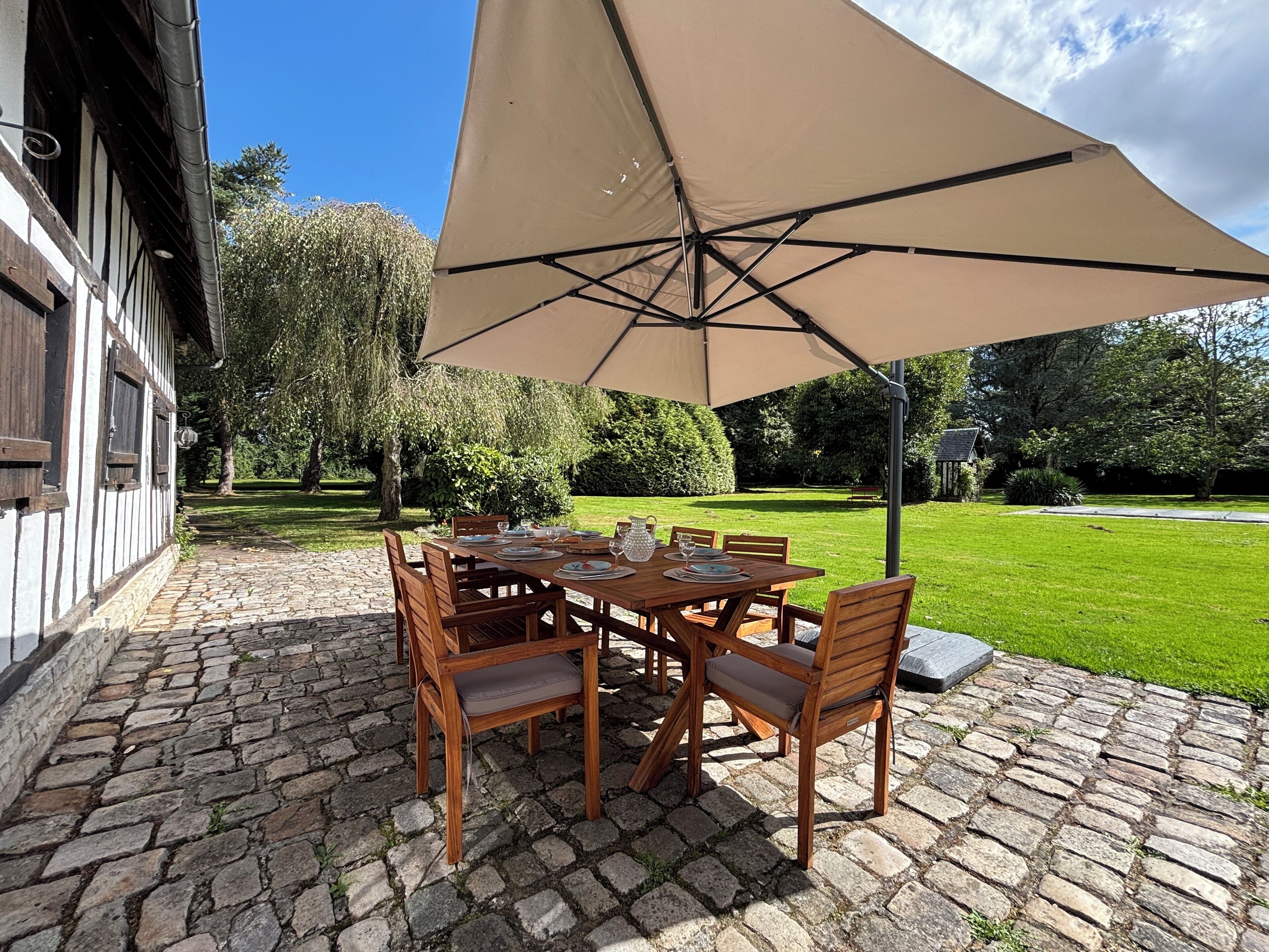 Une terrasse extérieure avec un sol carrelé en pierre, une table en bois dressée pour le repas et un grand parasol beige offrant de l’ombre.