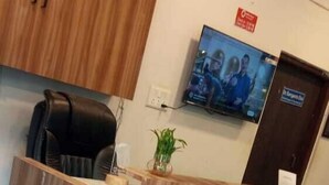 Un mostrador de recepción con acabado de madera, una silla de oficina negra, una planta en maceta y un televisor montado en la pared.