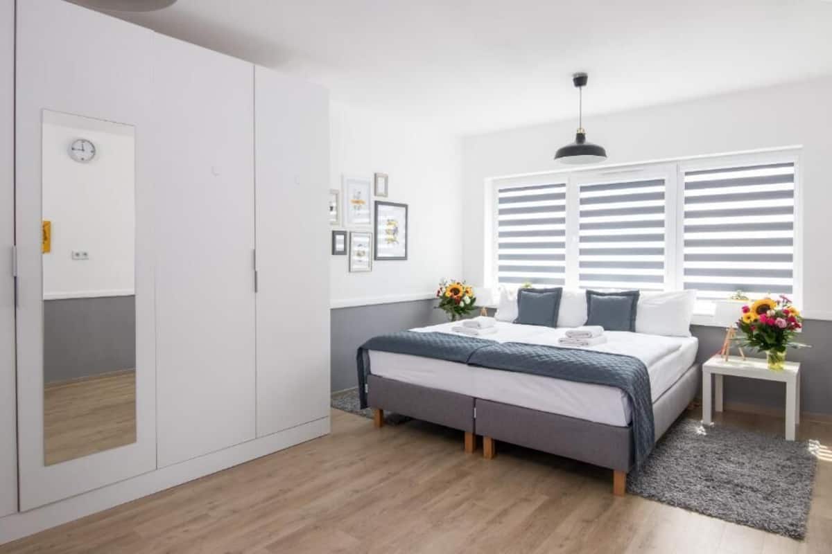A bedroom with a bed, bedside table, window with blinds, wardrobe, and a clock on the wall.