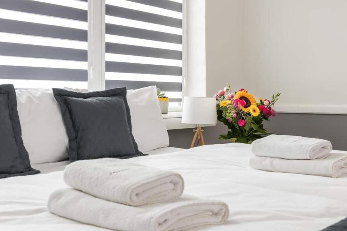 A neatly made bed with white linens and rolled towels, a vase of flowers, and a lamp on a bedside table.