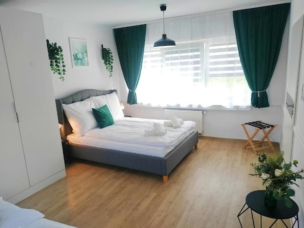 Un dormitorio con cama, ventana con cortinas, radiador, una mesita con planta y una silla.