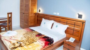 Quarto com cama de madeira, almofadas brancas, manta com estampa de leopardo, guarda-roupa de madeira, mesa de cabeceira com telefone e uma cadeira.