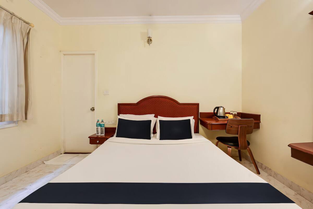 A hotel room with a large bed, a wooden nightstand with a lamp, a chair, and a small table with a kettle.