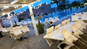 An outdoor seating area with plastic chairs and tables, a pool in the background, and string lights overhead. - Blu Apartments (Pietra Ligure)