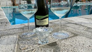 A bottle of Prosecco and two glasses of white wine on a stone table by a pool with a clear blue sky and palm trees in the background. - Blu Apartments (Pietra Ligure)