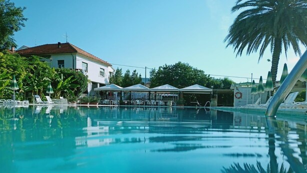 A clear blue swimming pool with lounge chairs and umbrellas, surrounded by greenery and a building with a red roof. - Blu Apartments (Pietra Ligure)