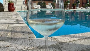 A clear glass of white wine on a stone surface near a pool with a reflection of buildings and palm trees. - Blu Apartments (Pietra Ligure)