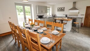 A modern kitchen with a dining area, featuring a central island, wooden chairs, and a view of the outdoors. - Segrwyd Uchaf (Denbigh)
