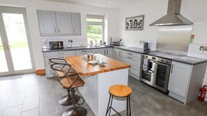 A modern kitchen with a central island, bar stools, stainless steel appliances, and a large window. - Segrwyd Uchaf (Denbigh)