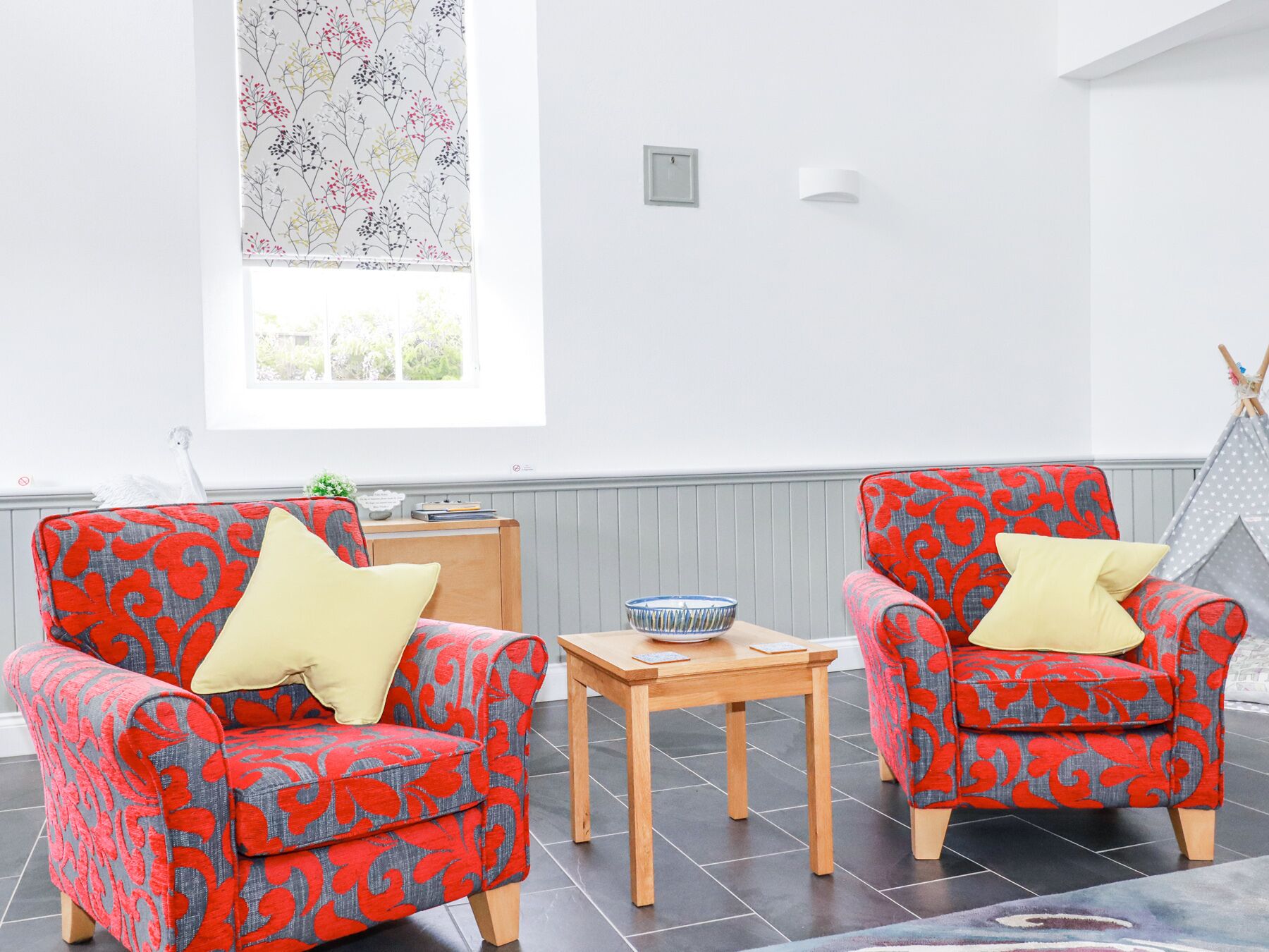 A room with two armchairs with red and grey floral upholstery, a small wooden table with a bowl, and a window with a floral view.