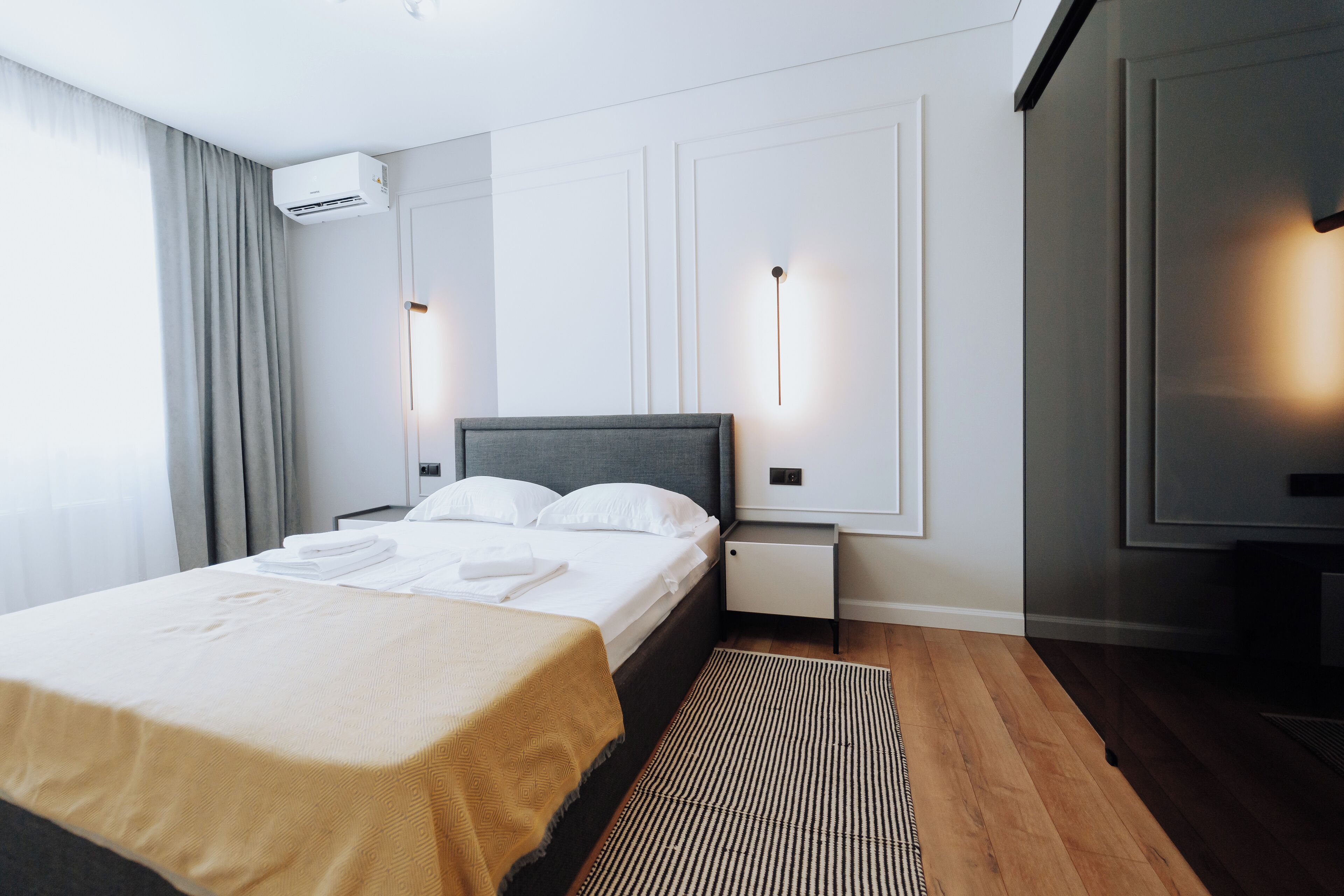 A modern hotel room with a large bed, a bedside table, an air conditioning unit, and a wall-mounted light fixture.