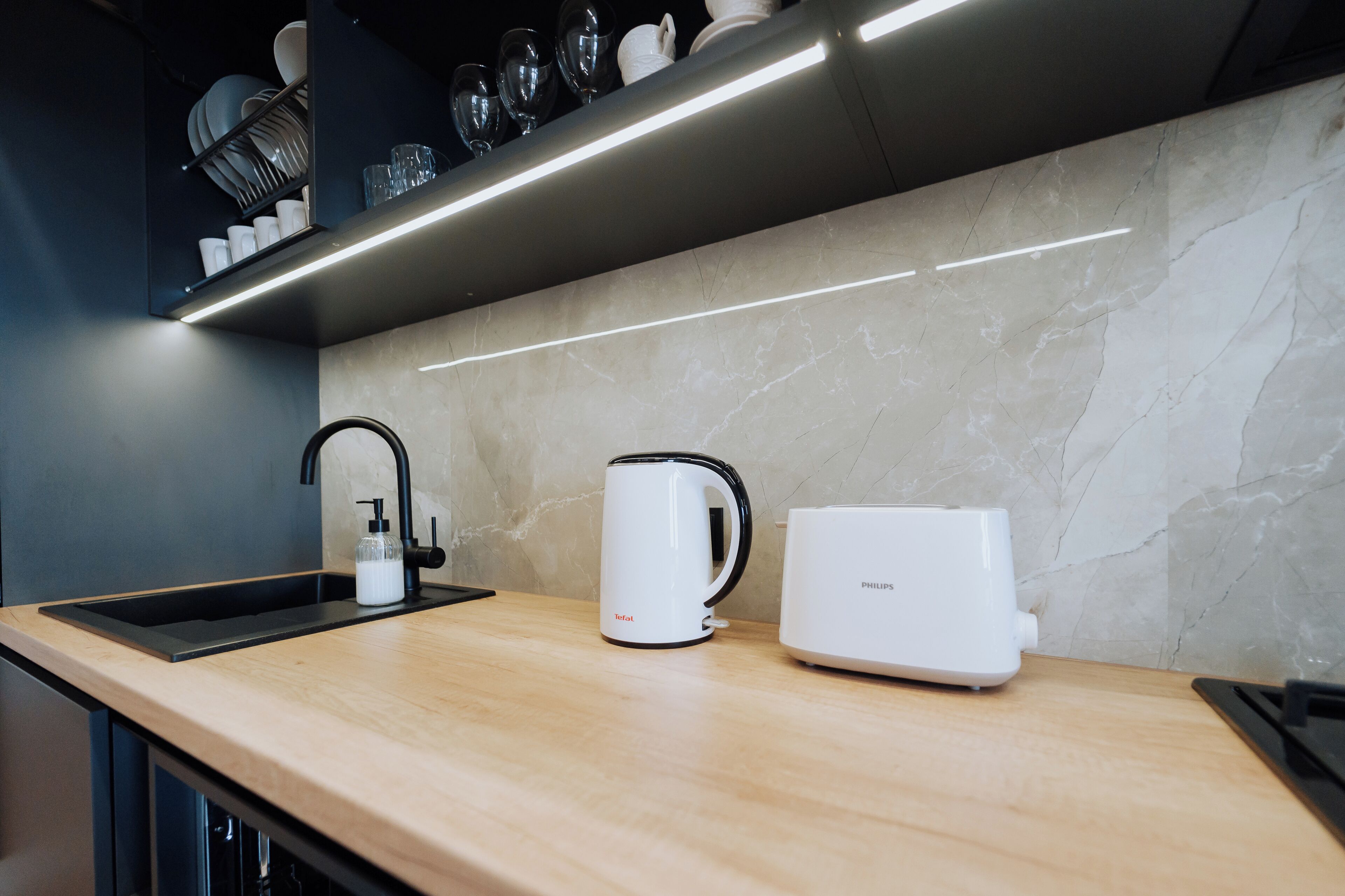 Una cocina moderna con un lavamanos negro, un hervidor blanco y una cafetera blanca sobre una encimera de madera.
