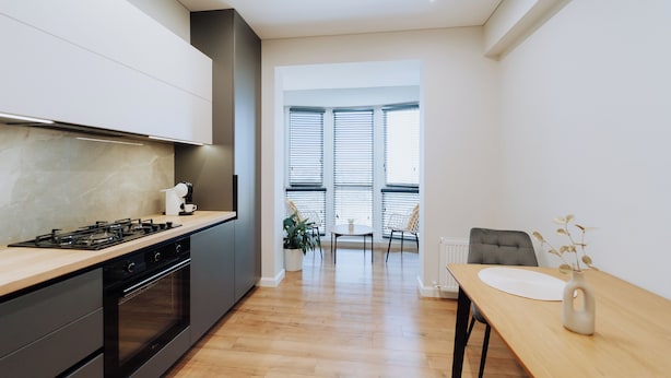 A modern kitchen with a gas stove, oven, and white cabinets. There is a dining area with a table and chairs, and a view of the outdoors through large windows.
